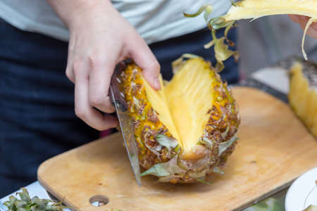 Hands of a man with a knife cut a juicy pineapple on a wooden board on the table around a dark background. Useful ingredientの写真素材