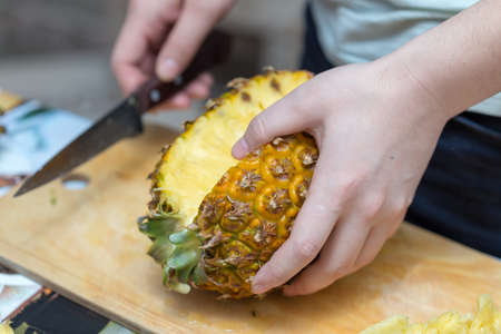 Hands of a man with a knife cut a juicy pineapple on a wooden board on the table around a dark background. Useful ingredientの写真素材