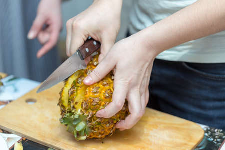 Hands of a man with a knife cut a juicy pineapple on a wooden board on the table around a dark background. Useful ingredientの写真素材