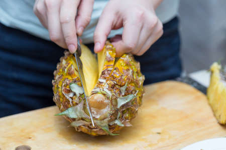 Hands of a man with a knife cut a juicy pineapple on a wooden board on the table around a dark background. Useful ingredientの写真素材
