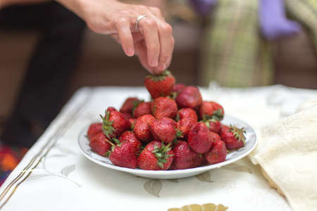 Sweet fresh strawberries on a plate on the table for, hands take and keep ripe red strawberries, a bunch of fresh strawberries in a ceramic bowl on a white table backgroundの写真素材