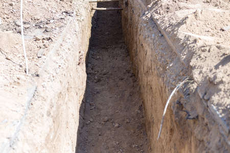 Laying pipelines of hot and cold water. Gas pipes are dug out by a tractor. Pit for urban communications. Excavations. Preparing for the construction of a house for residentsの写真素材