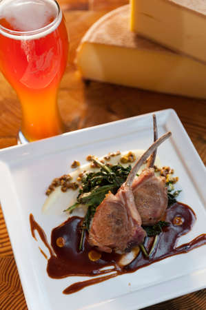 Grilled lamb chops on a white plate with a glass of beer and a block of round cheese.