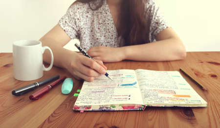 Young student girl writing in a colorful notebook with pens and marker around and a cup of coffee on...