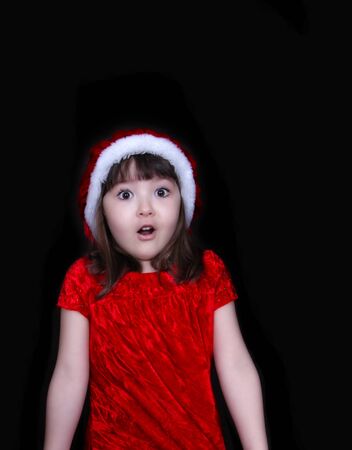sweet little girl in christmas dress and hat making a surprise face. isolated on blackの写真素材