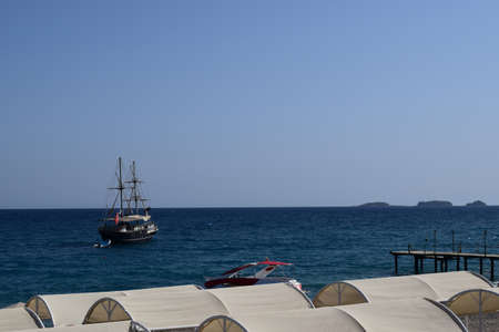 Old wooden sailing ship in sea, blue water. View on horizon from empty public beach with sunshades Small jetty boat is anchored close to the beach. Mediterranean Seaのeditorial素材