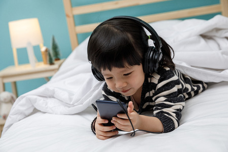 Cute little girl sing a song with smartphone in her bedroom, Happy asian child little girl listening the music with headphone on the bed, technology conceptの写真素材