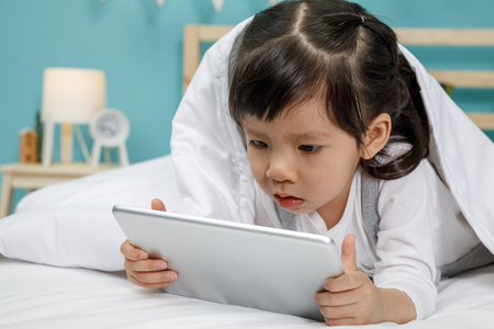 Child girl playing tablet in the blanket on the wooden bed in her bedroom, Happy asian child little girl using computer, learning concept at homeの写真素材