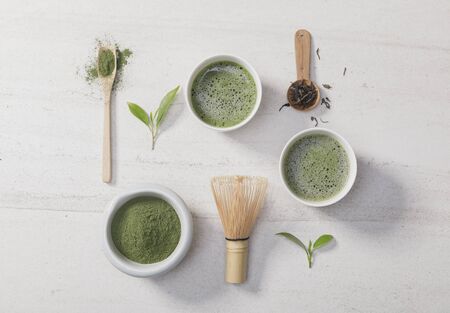 Organic matcha green tea powder in bowl with wire whisk and green tea leaf on white stone table,...
