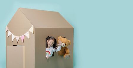 Asian little child girl playing with cardboard house with her teddy bear  isolated on blue long banner with copy space for your text, Creative at home with family conceptの写真素材
