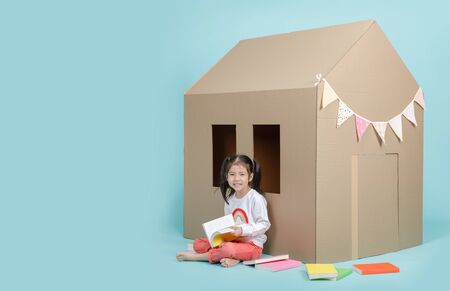 Asian little child girl reading a book with cardboard house isolated on blue long banner with copy space for your text. Homeschooling conceptの写真素材