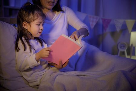 Mom reading a book with her daughter on bed in a dark bedroom at night before sleep, Parenthood and happy moments conceptの写真素材