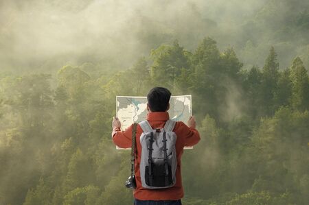 A young traveler man on the top of mountain with backpack in the green forest and look searching direction on location map, Adventure and travel in the mountains region conceptの写真素材