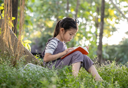 A pretty little Asian girl reading a book under the big tree.の写真素材
