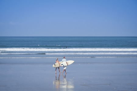surfer couple carrying surfboards walking on kuta beach bali with denpasar behindの写真素材