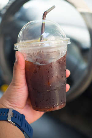 Man hand holding a cup of iced black coffee with steering wheel background (traveling concept)