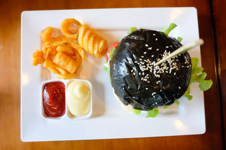 Black burger with meat pork, cheese, tomatoes, mayonnaise, french fries in a white plate.の写真素材