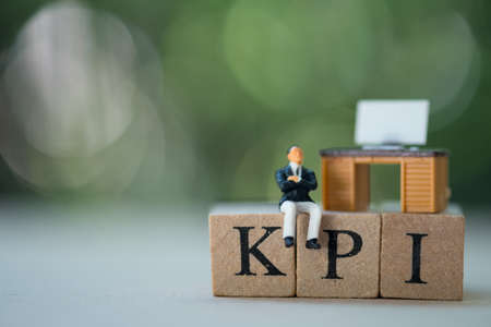 Miniature businessman sitting on kpi letters by wooden block in concept of key performance...