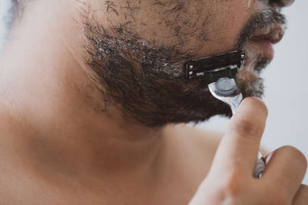 Young man with foam soap on his cheeks is standing and removing mustache by shaving from his face.の写真素材