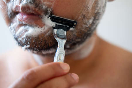 Young man with foam soap on his cheeks is standing and removing mustache by shaving from his face.