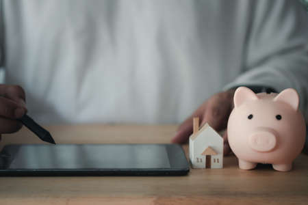 Young man use digital pen on tablet with miniature home and pink piggy bank on wood tableの写真素材
