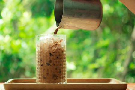 Pouring iced Coffee into ice in glass on wood tray with nature background