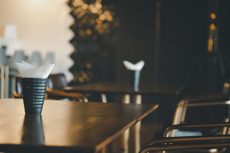 Close up of white paper tissue napkin in cup on wooden table. Restaurant tables waiting for customersの写真素材