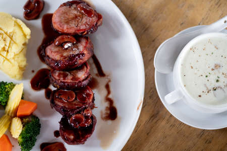 Pork fillet wrapped in bacon served with boiled vegetable and mushroom soup on wood tableの写真素材