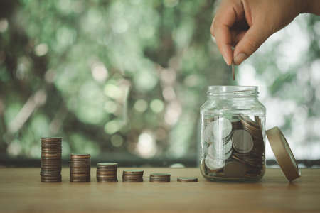 Hand holding coin put into money saving bottle on wood table against nature background.  Money saving idea to invest in the future.の写真素材