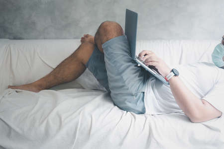 Young man wearing hygienic mask to prevent infection relaxing with a computer laptop seated on sofa in home. The concept of prevent infection, 2019-nCoV or coronavirusの写真素材