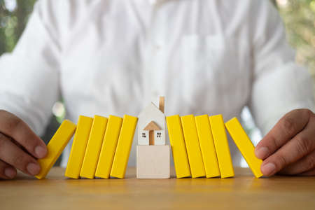 Close up of a human hand stop yellow dominoes fall to house wooden model. Real estate insurance. Management of risks. Security. Private property protection. Home accident insurance conceptの写真素材