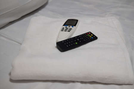 Air conditioner remote and TV remote places on white towel and white pillows in bed.の写真素材