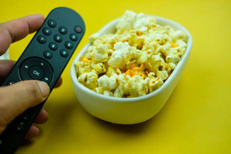 A hand use a TV remote control with a bowl of popcorn. The concept of watching TV at home