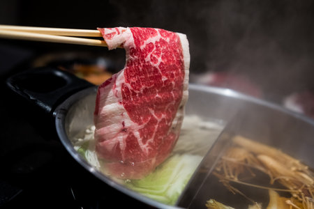 Hand using chopsticks pick premium raw beef sliced beef over the hot pot soup for Shabu Shabu or Sukiyaki dipping and dropping. Popular food is cooked piece by piece by the diner, served with dipping sauceの写真素材