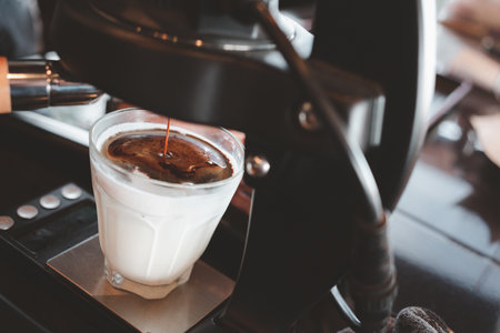 A clear glass cup of dirty coffee latte while the espresso machine maker is brewing and pouring hot and fresh coffee over cold fresh of milkの写真素材