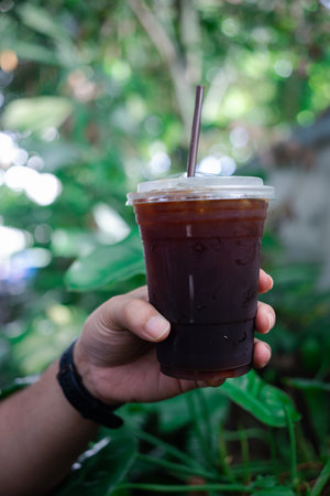 Hand holding a glass of iced black coffee with garden background in the morning.