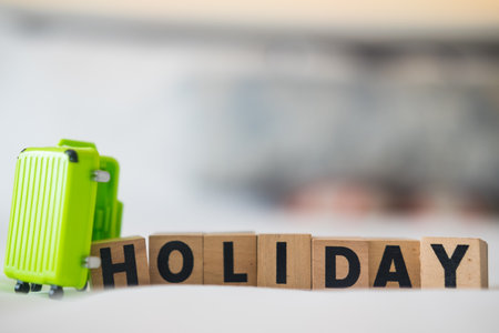 HOLIDAY word made with wood building blocks and green luggage on white bed.の写真素材