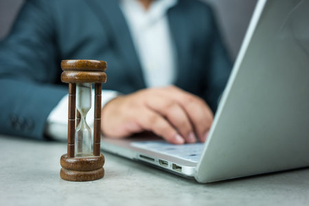 Businessman typing on a laptop keyboard next to a wood hourglass. Concept of time management,...
