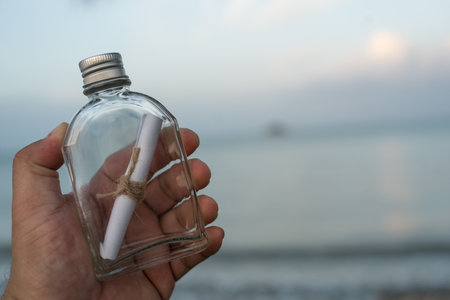 Paper letter in a glass bottle on hand over blurred sea water background. Crying for helpの写真素材