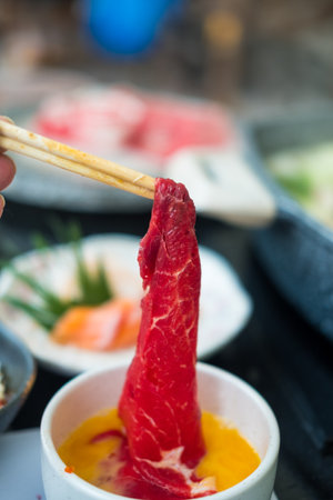 Close up of hand holding chopsticks with beef dip in raw beaten egg before boiling in hot potの写真素材