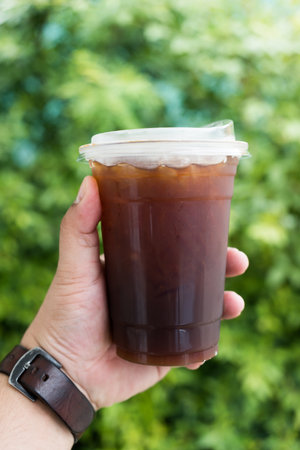 Man hand holding a plastic cup of iced americano or black coffee on the garden