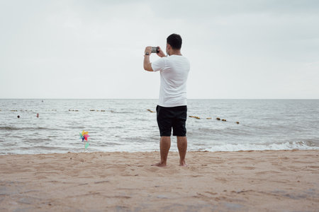 An Asian relaxing young man standing on the beach and using a smartphone takes a photo on the beachの写真素材