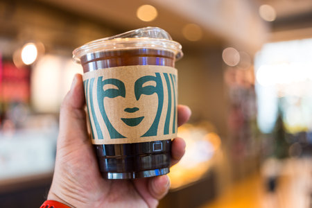 Bangkok, Thailand - November 23, 2023: Man holding a glass of iced Americano with a paper sleeve at Starbucks coffee shop.のeditorial素材