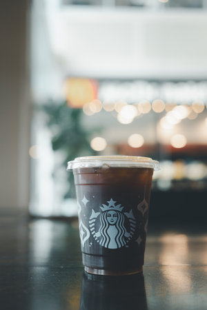 Bangkok, Thailand - December 7, 2023 : A takeaway plastic glass with iced Americano black coffee with lid on wooden table blurred lighting with copy spaceのeditorial素材