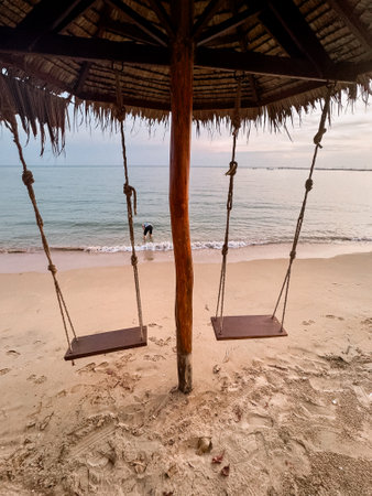 Two wood swings on the beach with a man walking into sea against the sunset on the beachの写真素材