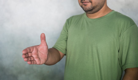 Friendly handsome bearded man wearing green T-shirt holding out his hand to side, for giving handshake on gray background with copy space. Smiling friendly offering handshake as greeting and welcomingの写真素材