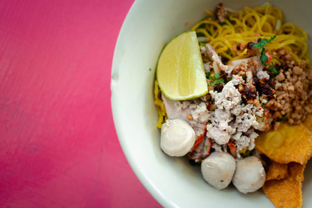 Egg noodles dried egg noodles with lime, minced pork, meatballs, and fried dumplings in white bowl....