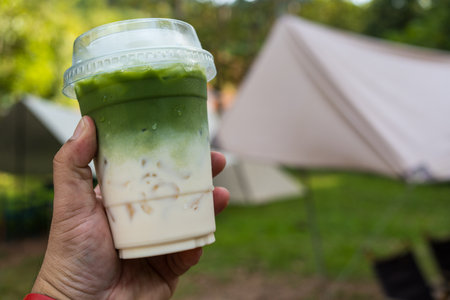 Cropped hand holding iced green tea matcha latte with camping backgroundの写真素材