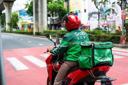 Bangkok, Thailand - August 14, 2024 : Grab delivery staff wearing green jacket and helmet holding, a motorbiker of Grab delivery service wait zebra crossing on street trafficのeditorial素材