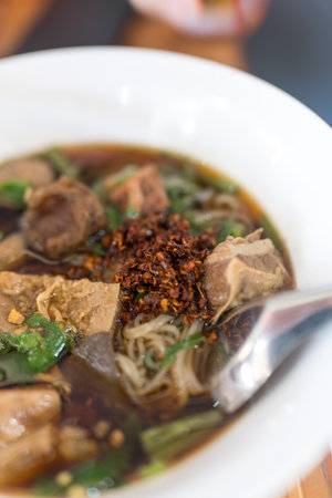 Beef noodle soup in a steaming bowl on a dark wooden table, packed with meat and veggies, suitable...
